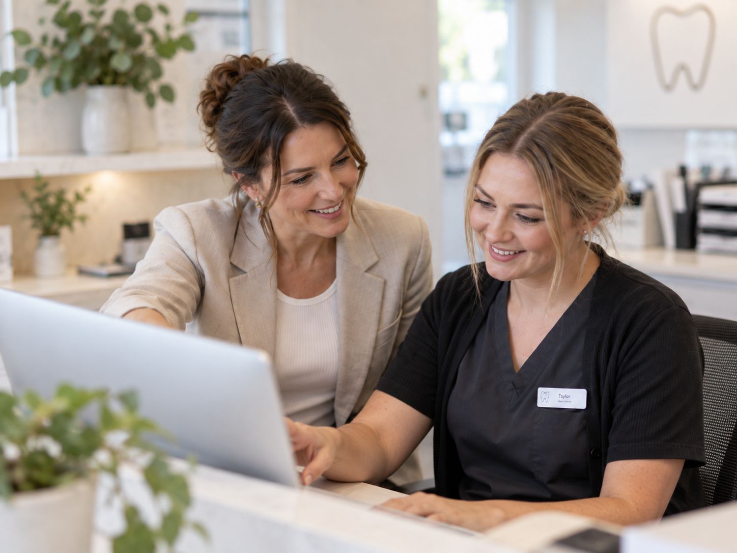 A consultant sitting beside a clinic receptionist, both smiling as they look at a laptop together